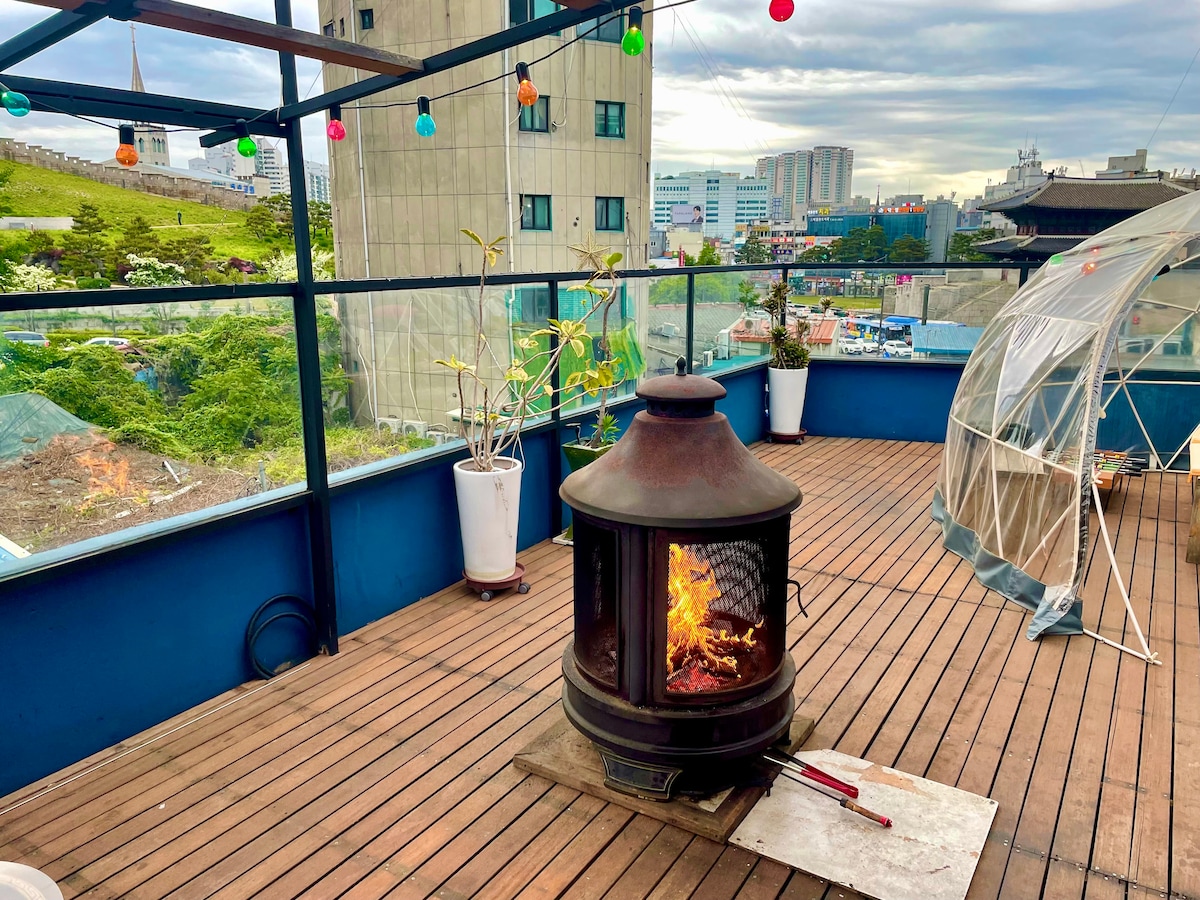 An outdoor terrace is shown with wooden flooring and a cozy outdoor dome structure. A fireplace is centrally placed, surrounded by decorative string lights. Potted plants are positioned around the space, creating a welcoming atmosphere for guests to enjoy.