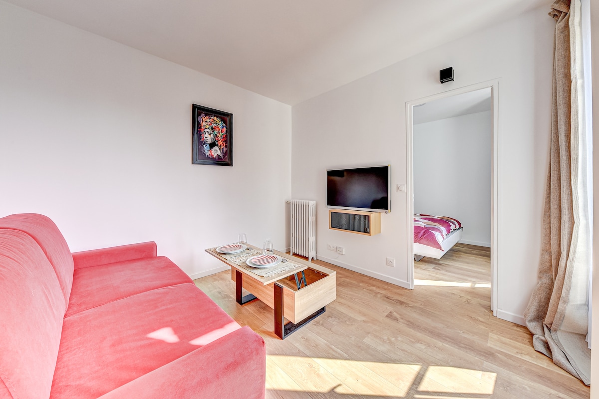 A bright living area features a red sofa and a wooden coffee table with decorative plates. A wall-mounted television is visible. Natural light illuminates the space, highlighting the light wood flooring. A doorway leads to the bedroom, and a piece of colorful wall art adds visual interest.