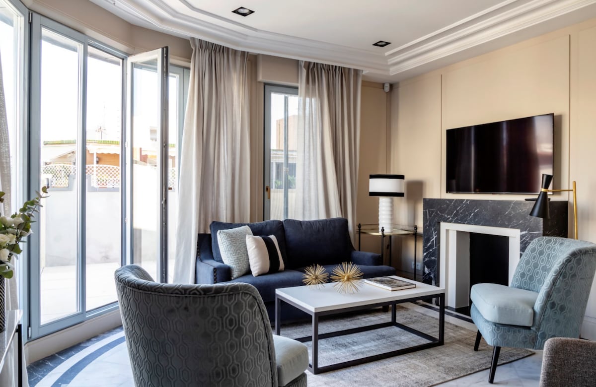 A stylish living area is showcased with large windows framed by light curtains. A comfortable blue sofa is positioned across from a sleek coffee table, complemented by two upholstered chairs. A wall-mounted television and a marble fireplace add a modern touch to the space.