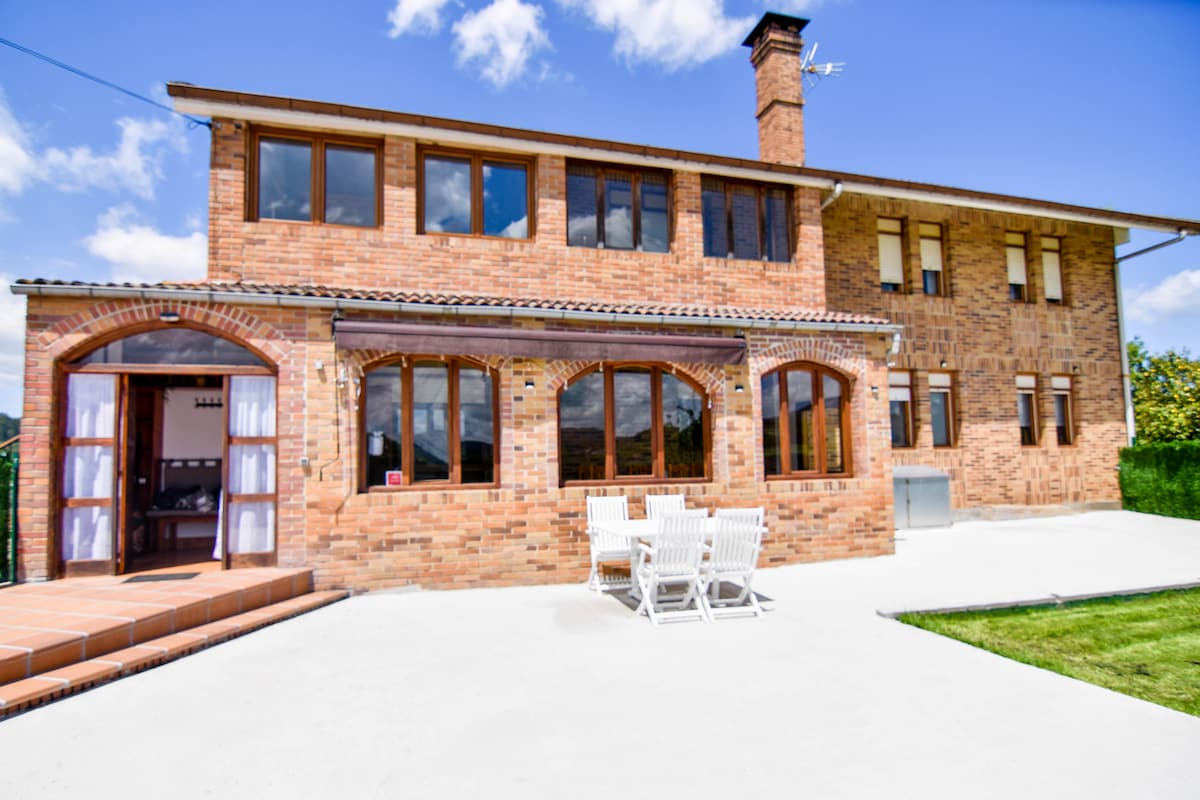A charming two-story brick house is shown with large windows allowing natural light to enter. A spacious patio is visible in front, with outdoor seating arranged around a table. The well-maintained garden adds greenery to the inviting exterior.