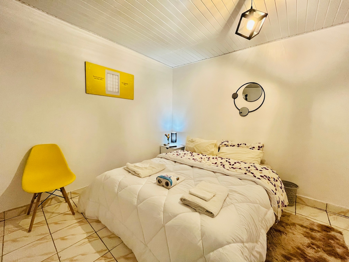 A cozy sleeping area features a spacious bed with a white quilt and neatly arranged towels. A light yellow accent chair adds a pop of color. Soft lighting from a ceiling fixture and a bedside lamp provides a warm ambiance. Art pieces adorn the walls, contributing to a welcoming atmosphere.