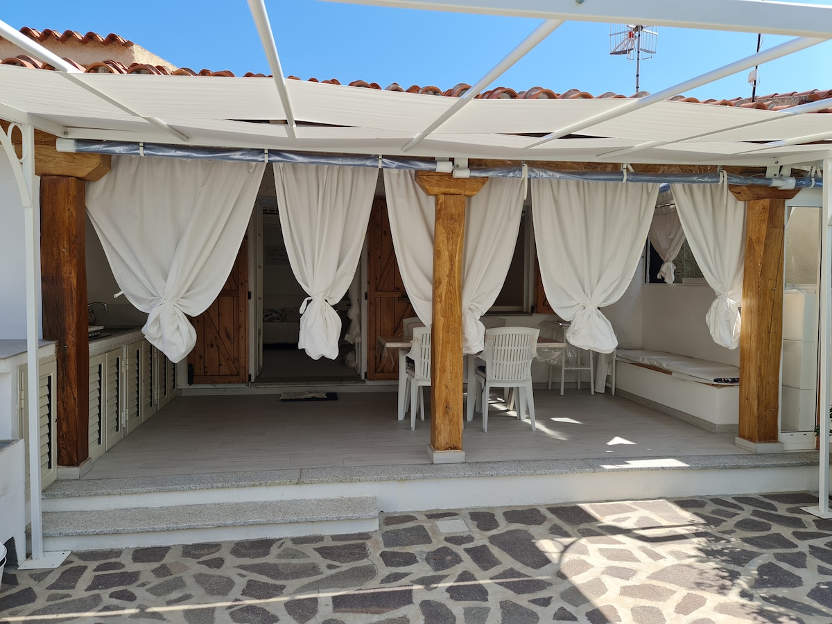 A covered verandah features a set of white outdoor chairs and a table, complemented by flowing white curtains. The stone flooring enhances the area, providing a spacious setting ideal for outdoor dining or relaxation.