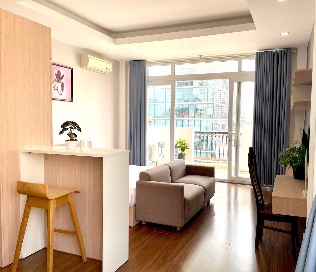 A bright, open living space showcases a seating area with a sofa and a small dining table. A balcony can be seen through large windows, allowing natural light to fill the room. Modern furnishings create a functional yet cozy atmosphere.