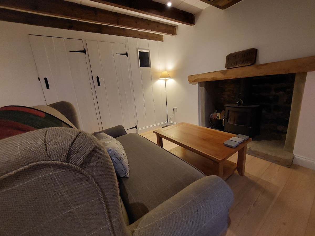 A cozy living room features exposed wooden beams and natural lighting. A gray sofa sits beside a wooden coffee table, while a stone fireplace with a wood-burning stove is positioned against the wall. A lamp provides additional soft illumination in the corner.