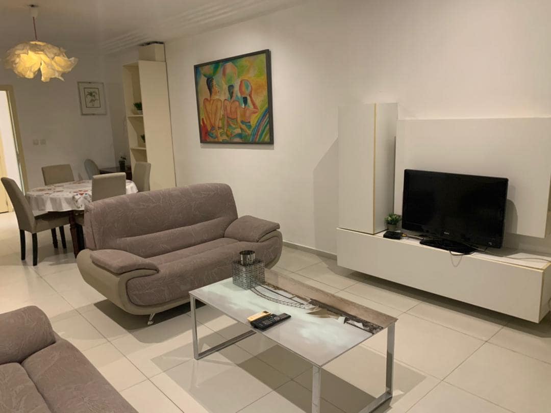 A modern living area features a soft gray sofa set and a sleek glass coffee table. A flat-screen television is positioned on a minimalist white console. Art decoratively hangs on the wall, while a dining area is visible in the background.