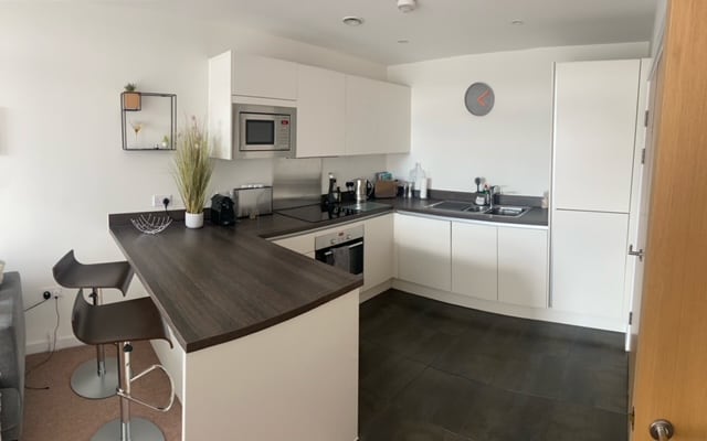 A modern kitchen area is presented, featuring sleek white cabinetry and a dark countertop. Appliances include a microwave and kettle, with a breakfast bar complemented by two stools. A wall clock and decorative plant add a touch of style to the contemporary space.