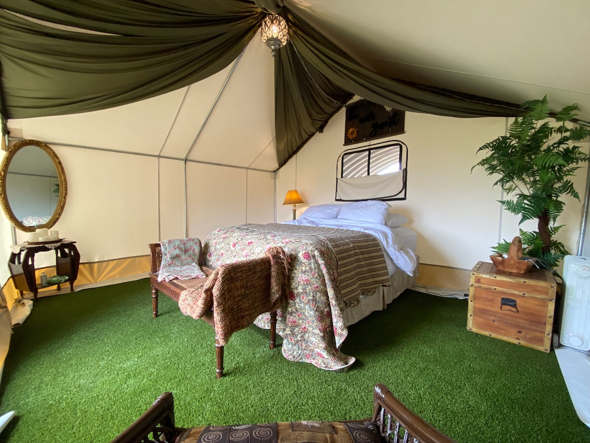 An inviting glamping tent features a spacious bed draped in soft linens. A patterned blanket is neatly folded at the foot. Plush seating is provided by a cozy bench and a decorative table. Green artificial grass covers the floor, enhancing the outdoor ambiance.