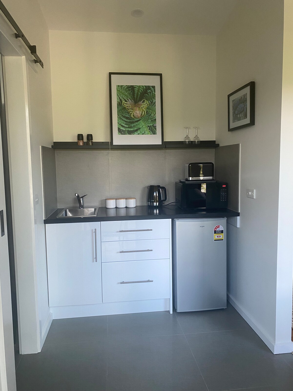 A compact kitchenette is shown, featuring a small refrigerator, a microwave, and a coffee maker on the countertop. A sink is located beneath a framed fern print, while white cabinets provide storage. A few glasses are neatly arranged nearby.
