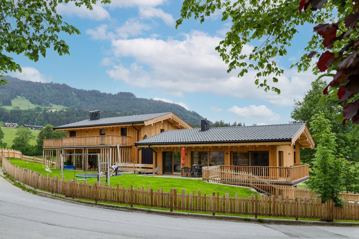The exterior of a chalet-style building is shown, featuring wooden construction and a sloped roof. A lush green lawn surrounds the property, which is framed by a wooden fence. Trees and mountains in the background complete the serene landscape.