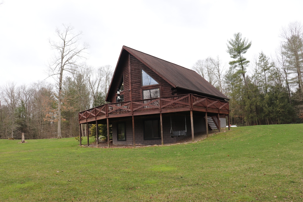 A spacious log chalet is featured, showcasing a wraparound deck with multiple seating areas. The structure is surrounded by an expansive lawn and a backdrop of trees, presenting a serene retreat in a natural setting.