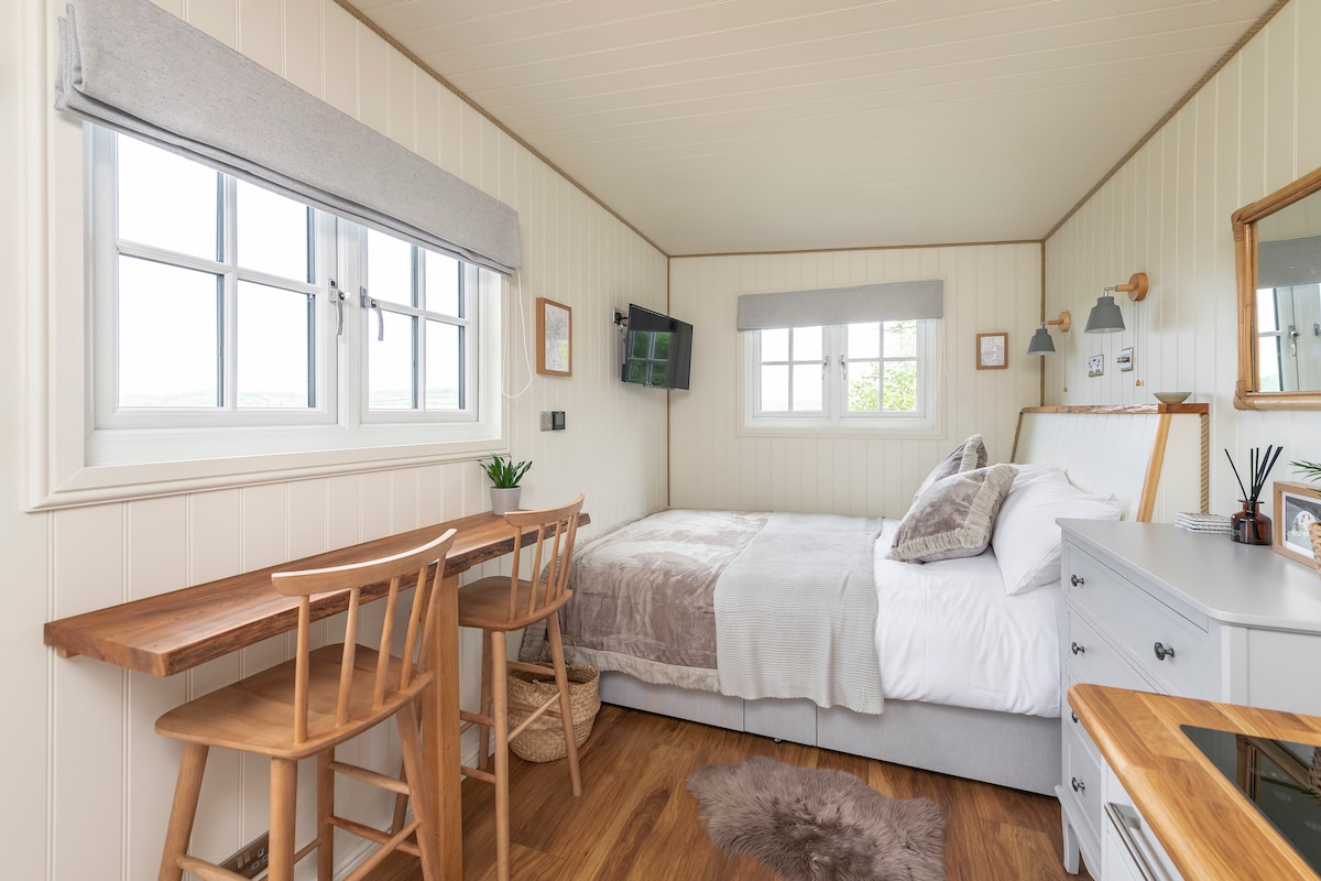 A compact lodge bedroom features a spacious bed with soft linens and a cozy throw. A small desk with two wooden chairs sits against one wall, while a mounted television and a mirror enhance the room's functionality. Natural light streams through the windows.