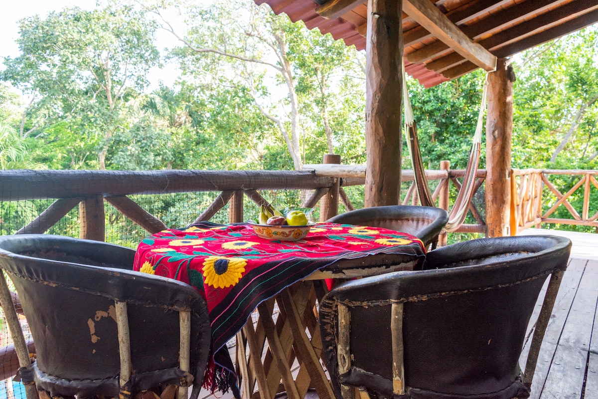 A wooden terrace is showcased, featuring a round table adorned with a vibrant traditional tablecloth. Surrounding chairs are positioned to create a cozy seating area. Lush greenery is visible in the background, enhancing the natural surroundings. A hammock can also be seen, inviting relaxation.