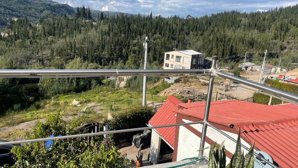 A metal railing frames the balcony view, revealing a landscape of greenery and distant hills. Below, traditional roof designs contrast with modern architecture, while a construction site is visible in the background, surrounded by natural elements.
