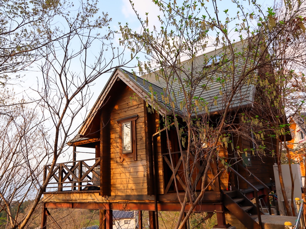 A wooden cabin is surrounded by trees, featuring a sloping roof and a balcony. The structure is elevated on stilts, allowing for views over the natural landscape. Soft evening light enhances the warm tones of the wood.