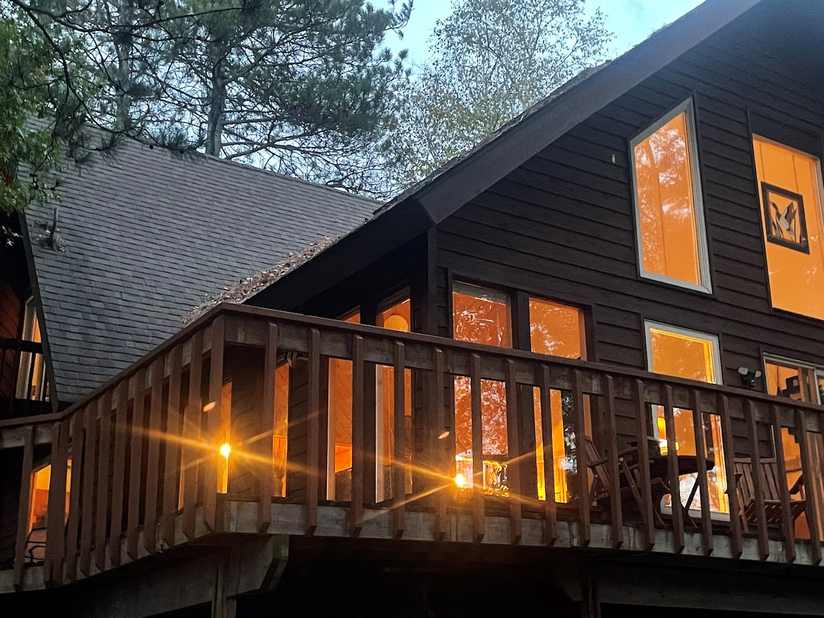 The exterior of the cabin highlights a spacious deck adorned with warm light, surrounded by trees. Large windows illuminate the interior, showcasing a welcoming atmosphere as the evening light filters through. The rustic architecture complements the natural setting.