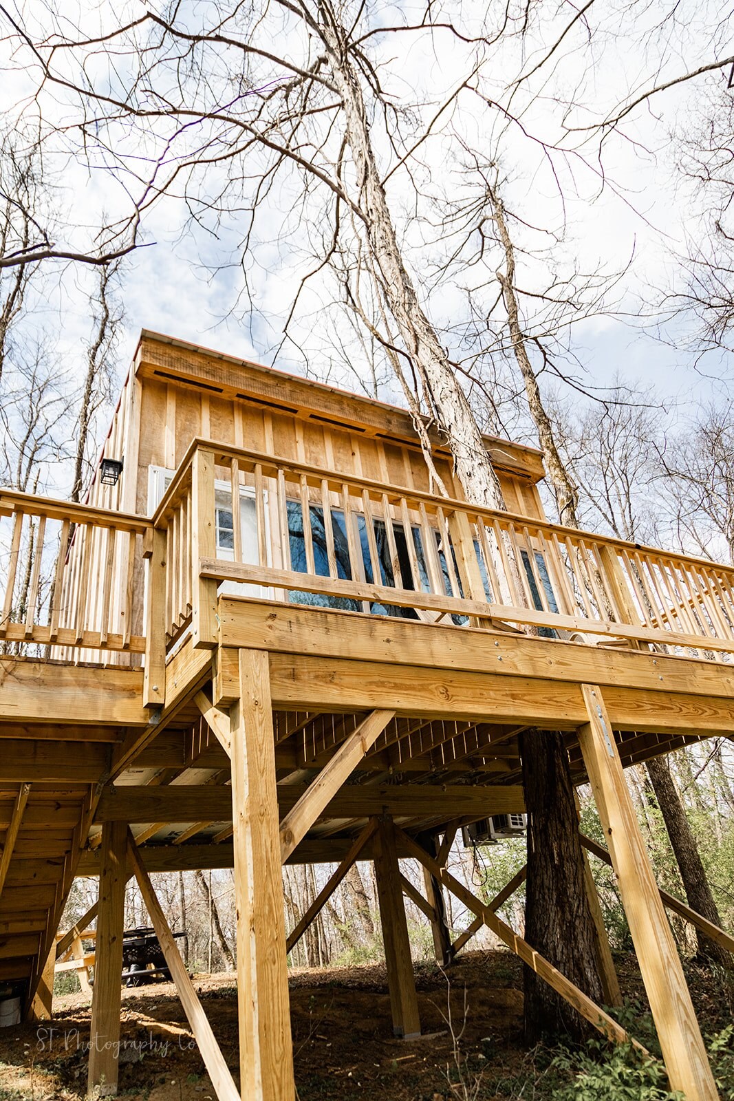 A rustic treehouse is perched above the ground, supported by wooden stilts. It features a wooden deck with a railing wrapping around, providing views of the surrounding trees. Large windows invite natural light into the space, showcasing its integration with the natural environment.