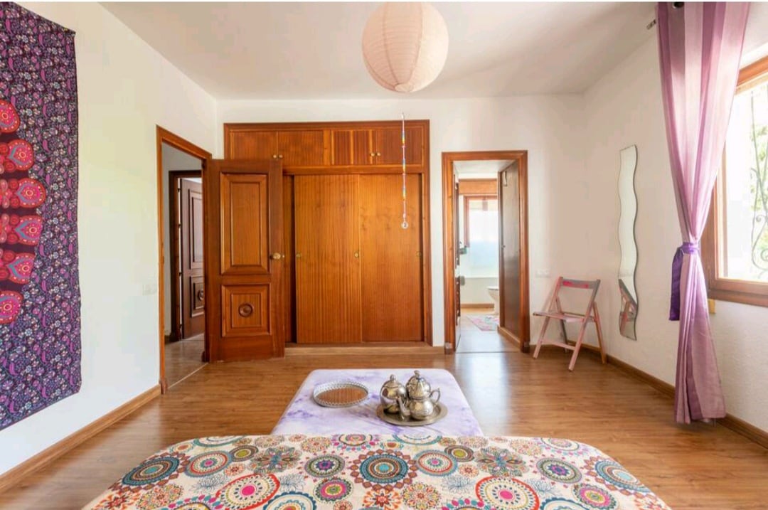 A spacious bedroom is highlighted by a colorful patterned bedspread and a large wall tapestry. Warm wooden cabinetry provides ample storage, and natural light fills the room through a window adorned with sheer curtains. A soft-toned mirror and a folding chair are visible in the background.