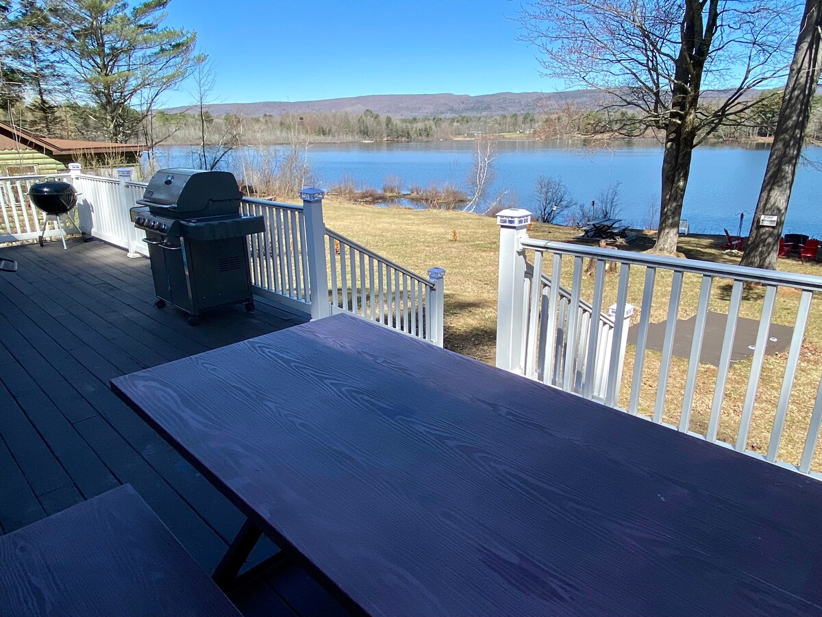 An outdoor deck features a picnic table overlooking the expansive lake. An attached gas grill stands nearby, surrounded by a serene landscape with clear blue water and trees in the background. The space invites relaxation and enjoyment of the natural surroundings.