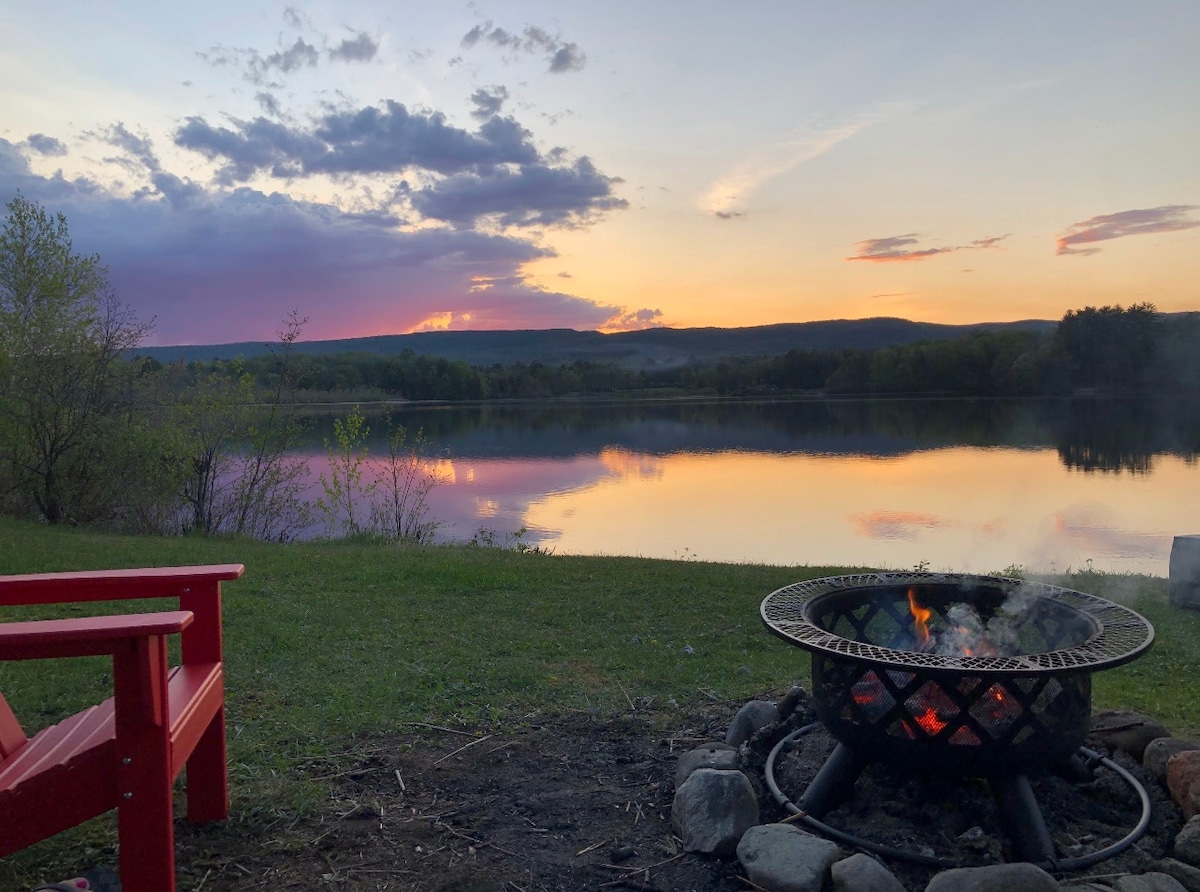 A tranquil lakeside scene features a firepit surrounded by stones, with flames flickering brightly. Red chairs offer seating, while the lake reflects the soft hues of the sunset, creating a serene atmosphere. Green grass extends towards the water's edge, framed by distant trees.