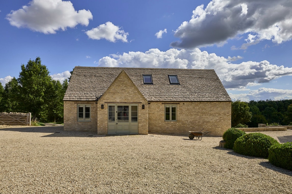 A newly built Cotswold stone cottage is showcased under a partly cloudy sky. The structure features a traditional design with a slate roof, two skylights, and large front doors. A gravel driveway leads to the entrance, framed by neatly trimmed bushes.