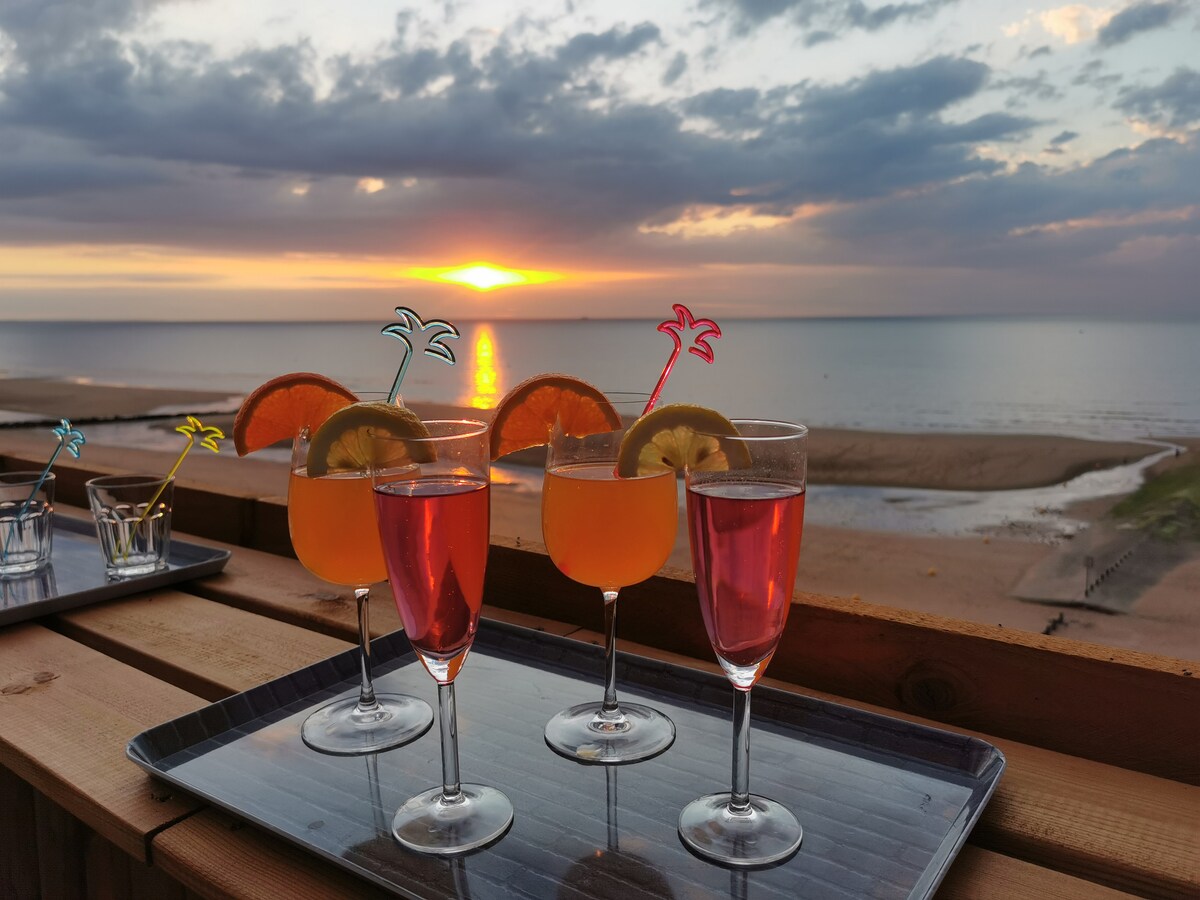 Four colorful cocktails are set on a wooden tray, with garnishes including orange and lemon slices. The backdrop features a stunning sunset over calm waters, casting soft hues of orange and pink across the sky, creating a serene atmosphere.