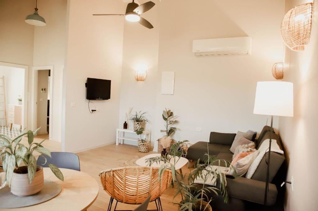 A well-lit living area features a comfortable sofa and a circular coffee table surrounded by plants. A wall-mounted television is positioned above a small console. A ceiling fan enhances the airy atmosphere, while light fixtures provide soft illumination.