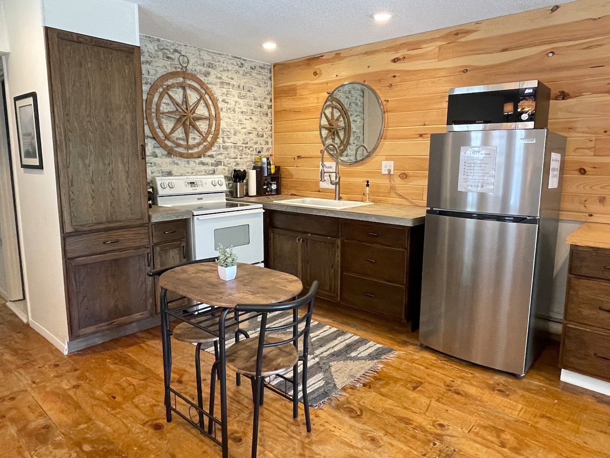 A functional kitchen area features wooden cabinetry and a modern stainless steel refrigerator. An electric stove and sink are positioned against a backdrop of stylish wood paneling and patterned wallpaper. A small round table with two chairs provides a cozy dining space.