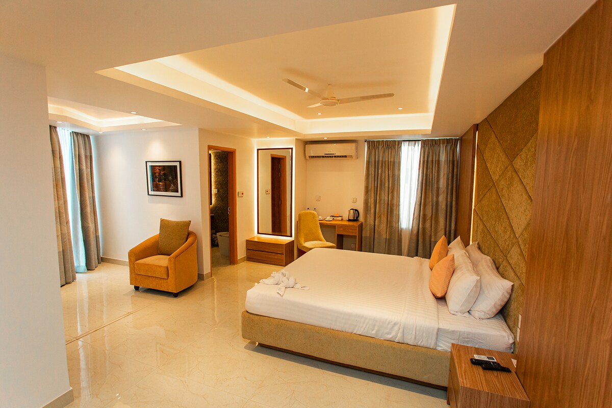 A spacious Deluxe Bedroom features a minimalistic design with soft lighting illuminating the textured wall behind the bed. A cozy chair is positioned near a window, while a vanity area is visible beside the bed, enhancing the room's functional layout.