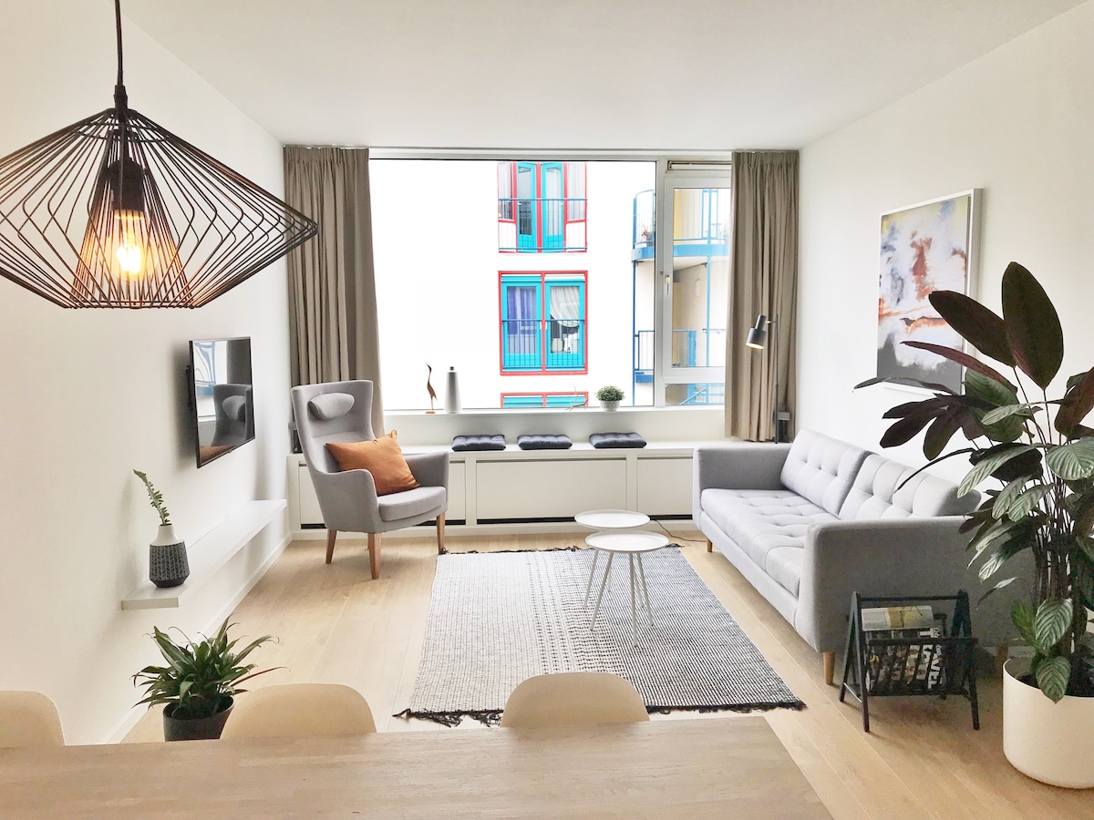 A light-filled living area showcases a modern gray sofa facing a flat-screen TV mounted on the wall. A stylish pendant light hangs above, with a cozy armchair nearby and a small coffee table positioned on a textured rug. Large window offers a view of colorful buildings.