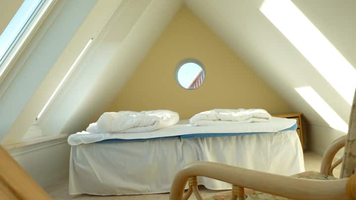 Dhoma e gjumit në loft, me perde të errëta në të gjitha shpatet dhe pamje të pjesës së vjetër të qendrës së qytetit të Løkken. 