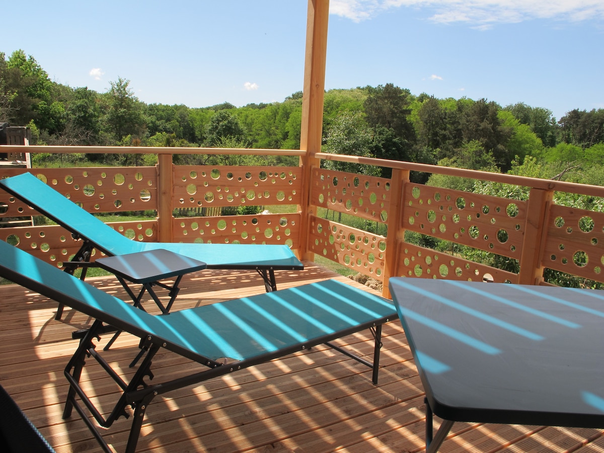 A private terrace is shown, featuring two lounge chairs positioned for relaxation. A small table complements the space, surrounded by a wooden railing with decorative cutouts. Lush greenery can be seen in the background, enhancing the serene atmosphere.
