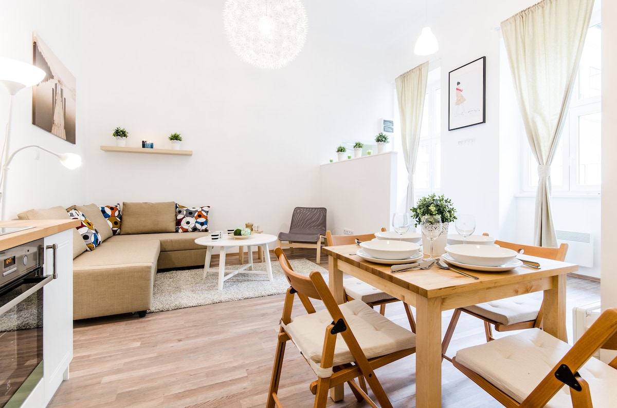 A light-filled living space features a neutral-colored sectional sofa with decorative cushions and a small shelf above. A wooden dining table is set for four, accompanied by foldable chairs. Floor-to-ceiling windows are adorned with light drapes, enhancing the airy ambiance.