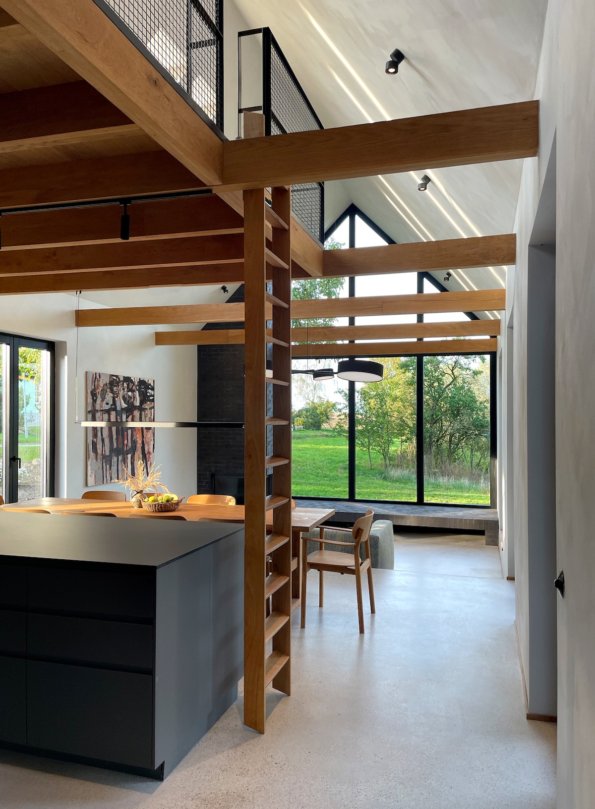 A spacious interior showcases a modern kitchen with a black island, paired with wooden shelves and a dining area. Large windows offer views of the outdoor greenery, while natural light enhances the wooden beams and contemporary design.