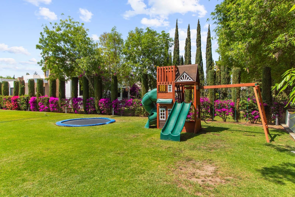 A spacious backyard features a wooden playset with a slide and climbing structure alongside a circular trampoline. Lush green grass surrounds the play area, while vibrant bougainvillea and tall cypress trees provide a colorful backdrop, creating an inviting outdoor space for relaxation and play.