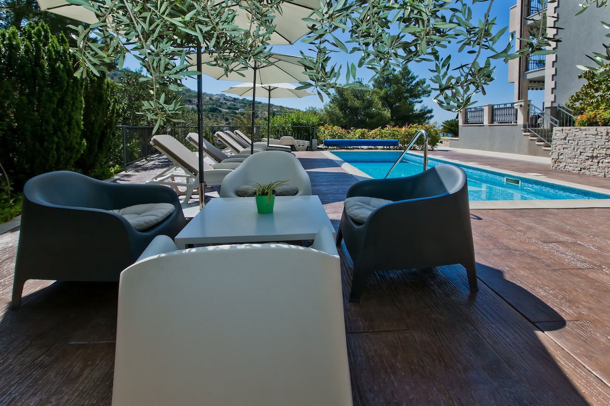 An outdoor area is featured, with a swimming pool surrounded by lounge chairs and shaded by umbrellas. Comfortable seating options include two modern chairs and a coffee table, creating a relaxed atmosphere for enjoying the warm weather.
