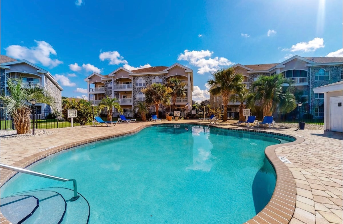 An outdoor pool is surrounded by palm trees and tropical landscaping, creating a calm setting. Lounge chairs are positioned around the pool area. The three-story condo building stands in the background, with a clear blue sky overhead, enhancing the inviting atmosphere.