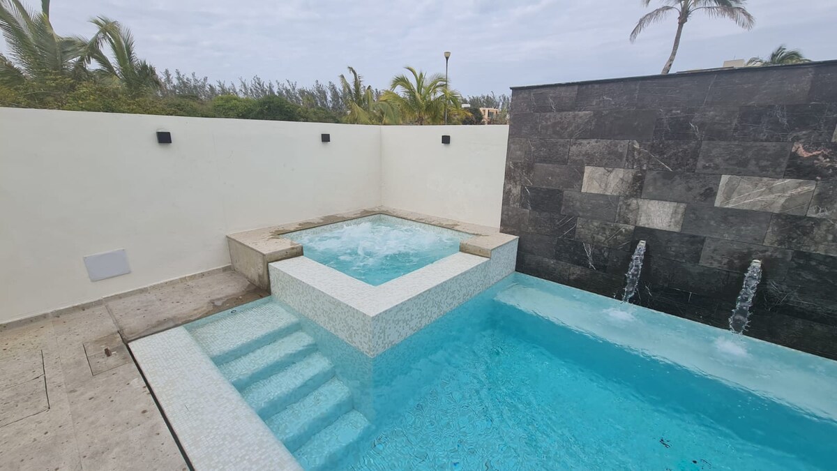 A private pool area is visible, featuring a spacious jacuzzi attached to the pool. A smooth stone wall with three water features complements the serene atmosphere. Surrounding palm trees contribute to the tropical feel of the outdoor space.
