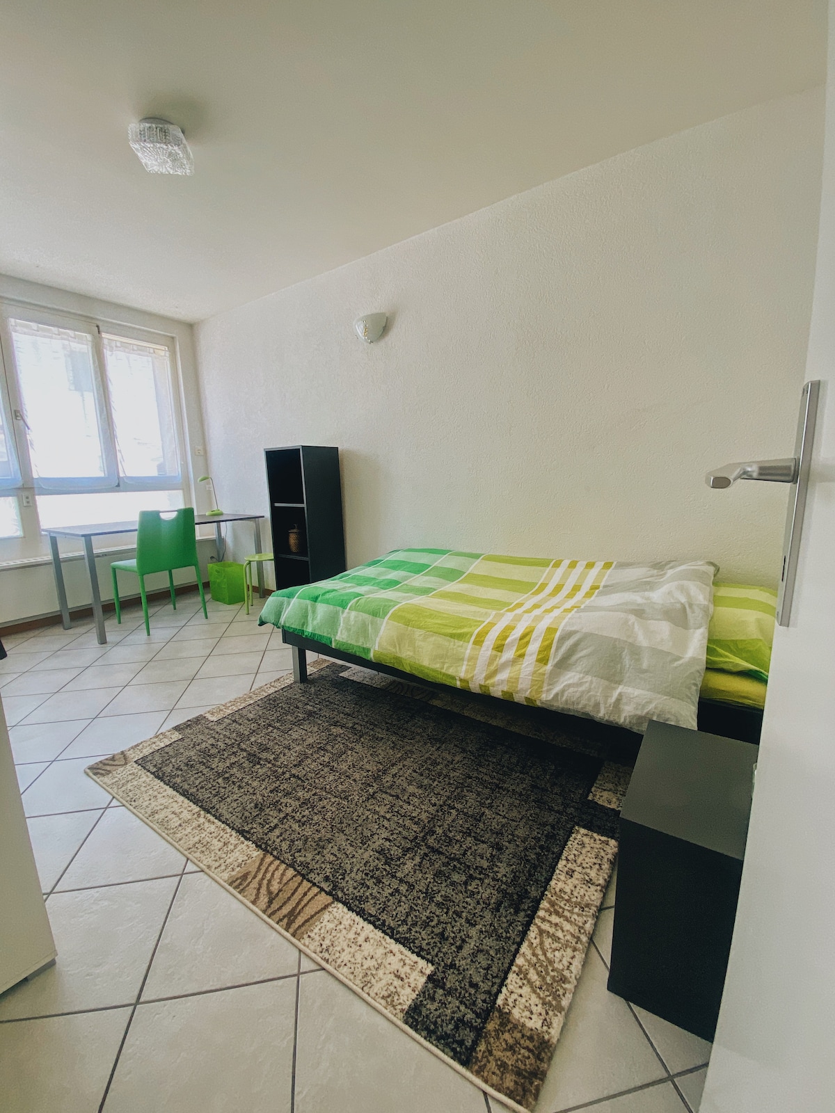 A compact studio room features a single bed covered with a green and white striped blanket. A small table and green chair are positioned by the window, allowing natural light to enter. A dark cabinet and a rug provide additional decor.
