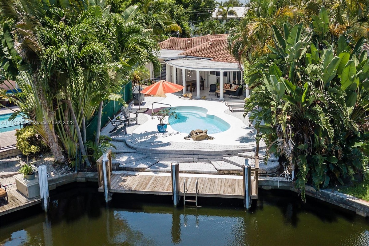 A waterfront property is shown, featuring a pool surrounded by palm trees and outdoor seating. An orange umbrella provides shade over a lounge chair. A private dock extends into the tranquil canal, underlining the home's serene outdoor living space.