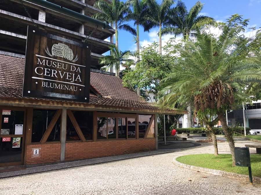 A brown brick building is shown with a large sign displaying 'Museu da Cerveja Blumenau' at the entrance. Lush palm trees and green foliage are visible around the structure, while a stone pathway leads toward the entrance. Clear blue skies add to the serene atmosphere.