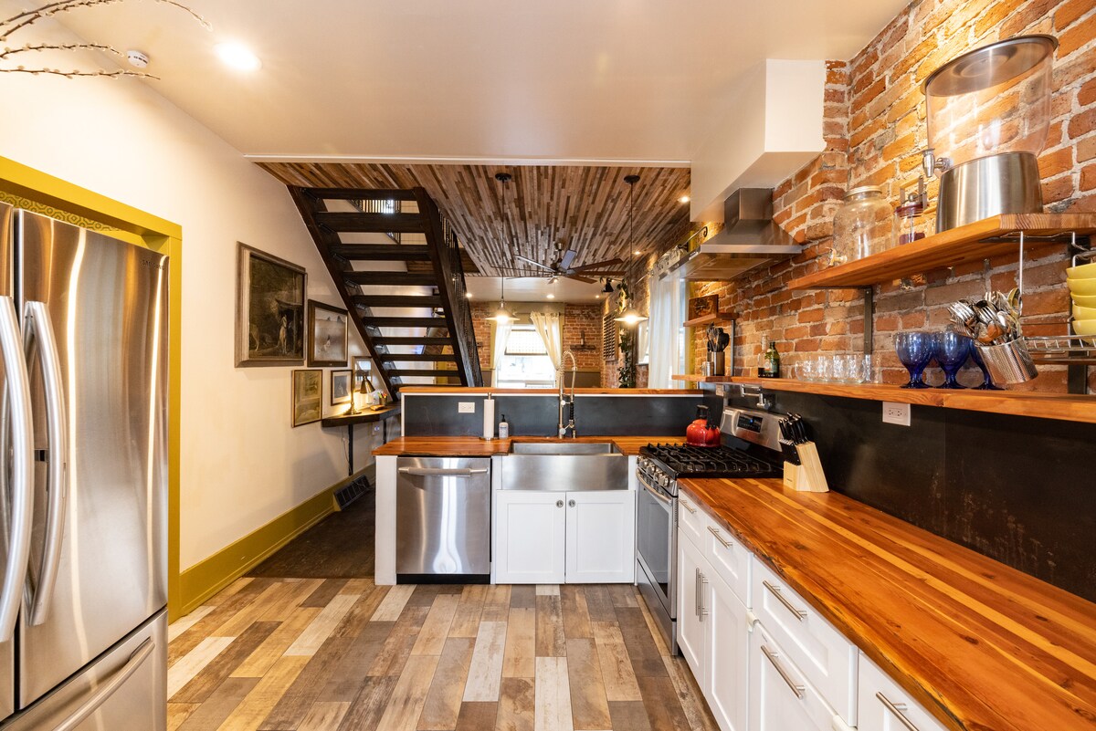 A modern kitchen features stainless steel appliances, including a large fridge and gas stove. Exposed brick walls add character, complemented by wooden countertops and open shelving displaying various kitchen items. Natural light streams in through nearby windows, enhancing the inviting space.