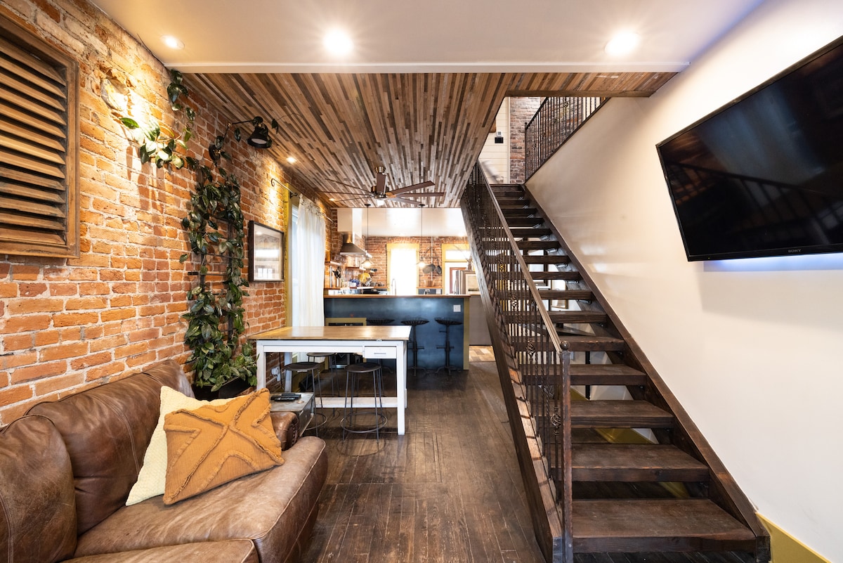 A stylish living area features exposed brick walls and a wooden ceiling with warm lighting. A comfortable leather couch is paired with a sleek dining table. A staircase ascends to the upper floor, leading to the kitchen area in the background.