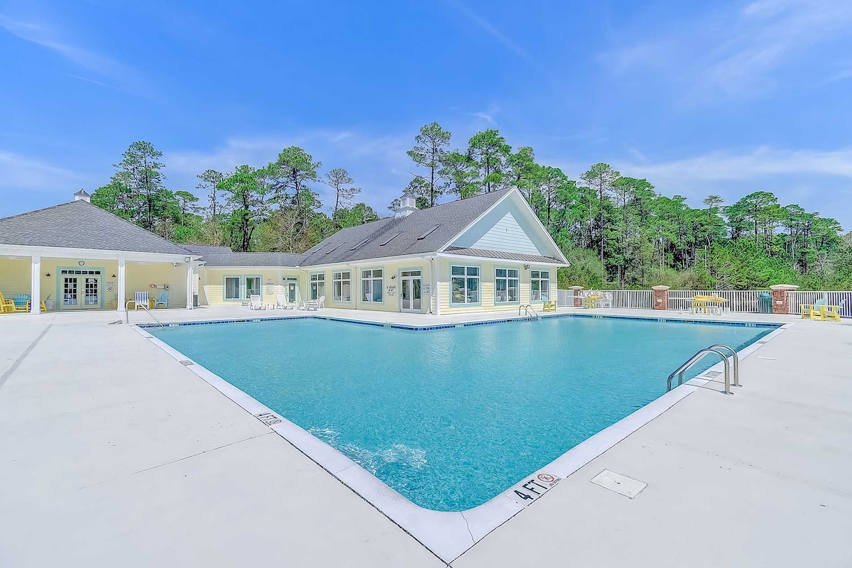 An outdoor pool is surrounded by a spacious deck and well-maintained landscaping. The building features large windows and a covered area nearby for lounging. The clear blue water reflects the sky, creating a serene environment with ample seating available around the pool.