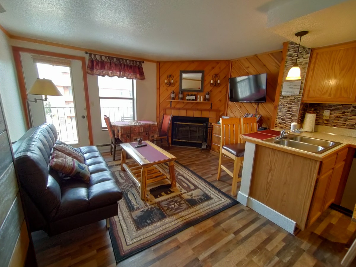 The spacious living area features a comfortable sofa with decorative cushions and a wooden coffee table. A dining table sits nearby, complemented by a bright window allowing natural light. The kitchen area includes wooden cabinets and a sink, while a fireplace adds warmth to the space.