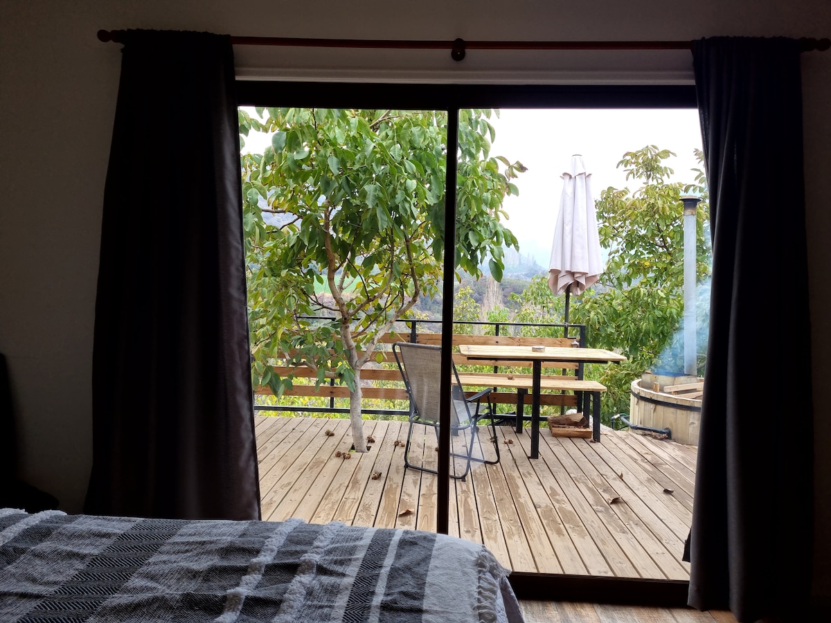 A spacious sliding door opens to a wooden deck featuring a dining table with chairs and an umbrella. Surrounding greenery offers a natural view, while a hot tub is positioned nearby. Light filters softly through the curtains in the interior space.
