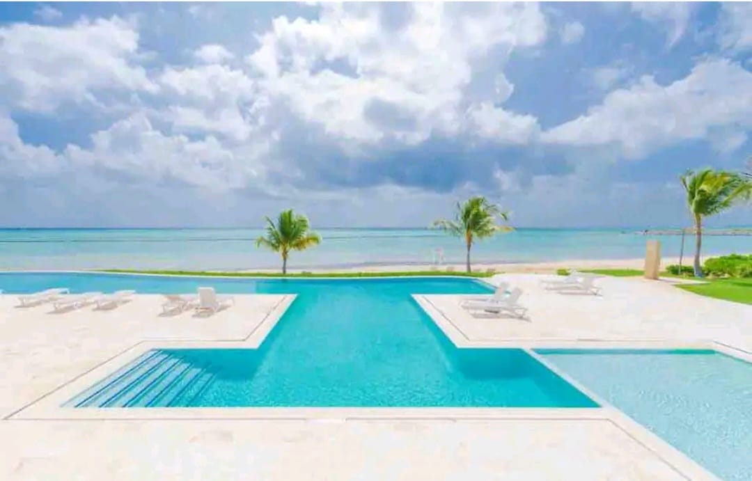 A spacious pool area is visible, featuring a large swimming pool with a shallow entry. The pool is surrounded by numerous lounge chairs, with palm trees lining the beachfront. The calm ocean water extends into the distance under a partly cloudy sky.