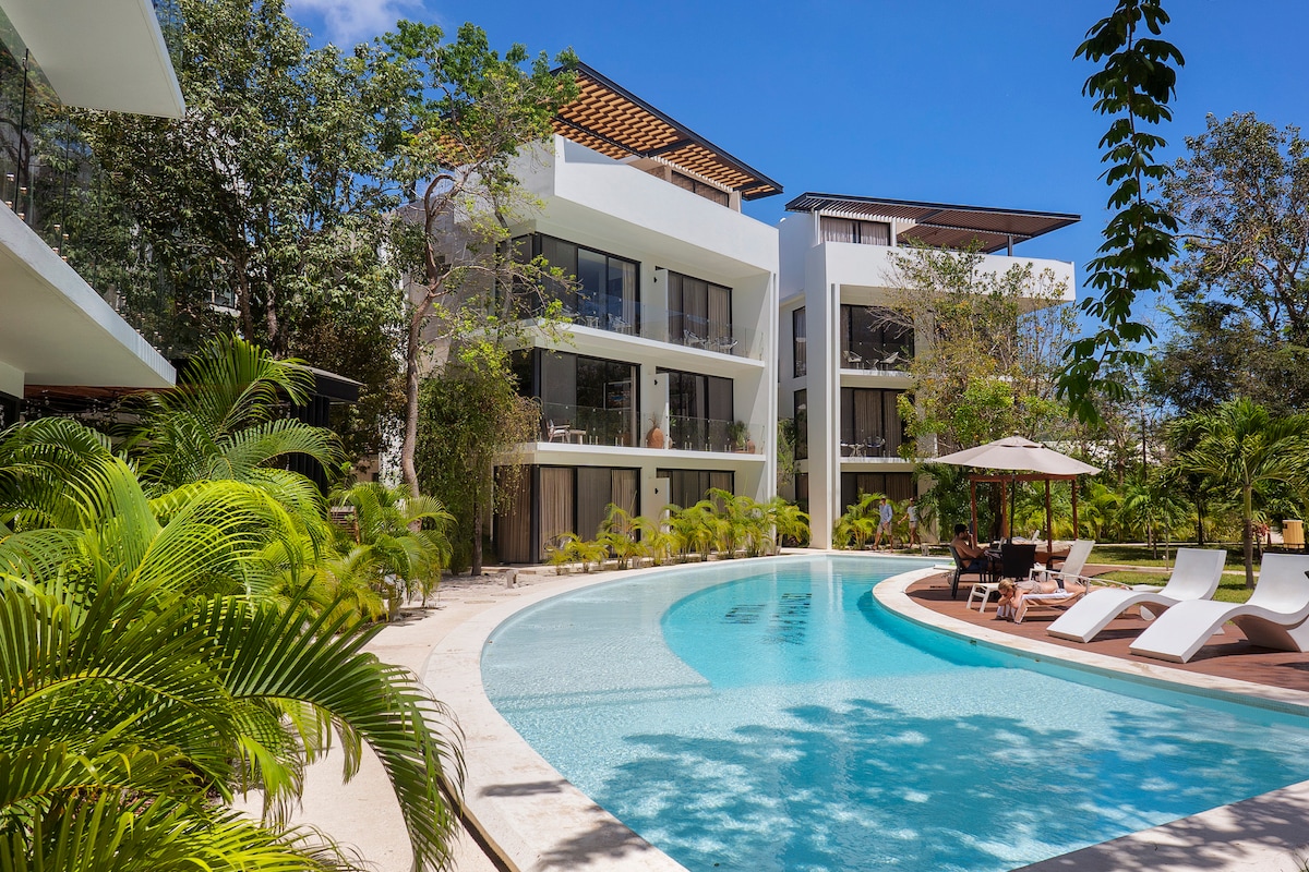 A modern luxury condo complex is surrounded by lush tropical landscaping. The inviting lap pool features a smooth, reflective surface and is bordered by loungers under shaded umbrellas. The clear blue sky complements the serene atmosphere of the outdoor space, promoting relaxation.