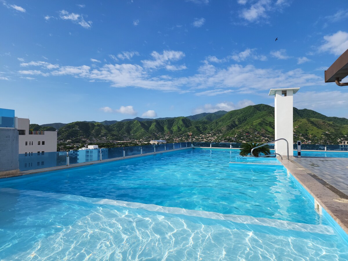 A rooftop infinity pool reflects the bright blue sky and surrounding green hills. The tranquil water appears inviting, framed by a clean stone border. A modern structure with a small diving area is visible at one end of the pool, creating a serene leisure space.