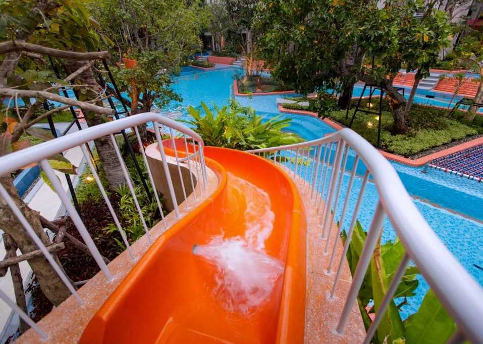An orange waterslide curves down into a serene pool area, surrounded by lush greenery and palm trees. The water flows from the slide into the inviting pool, which features various shades of blue. Relaxing lounge chairs are positioned along the edges of the pool.
