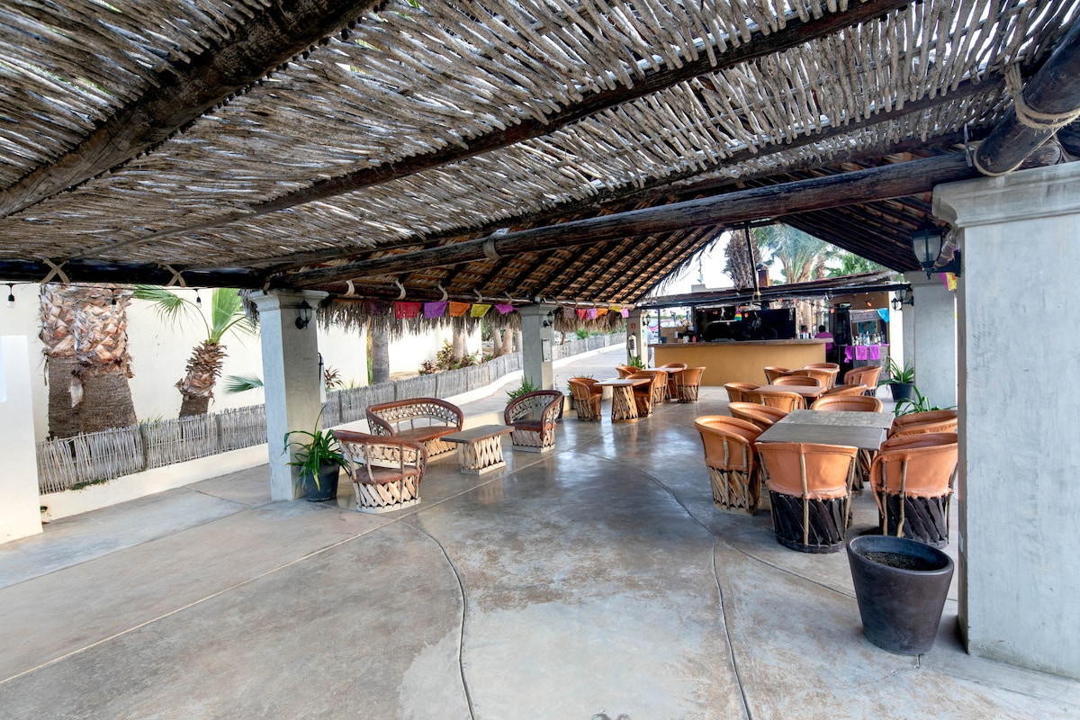 A spacious outdoor dining area features an open roof with natural thatch for shade. Wooden tables are surrounded by comfortable seating, creating a casual atmosphere. Potted plants add a touch of greenery, and a bar is visible in the background, enhancing the inviting space.